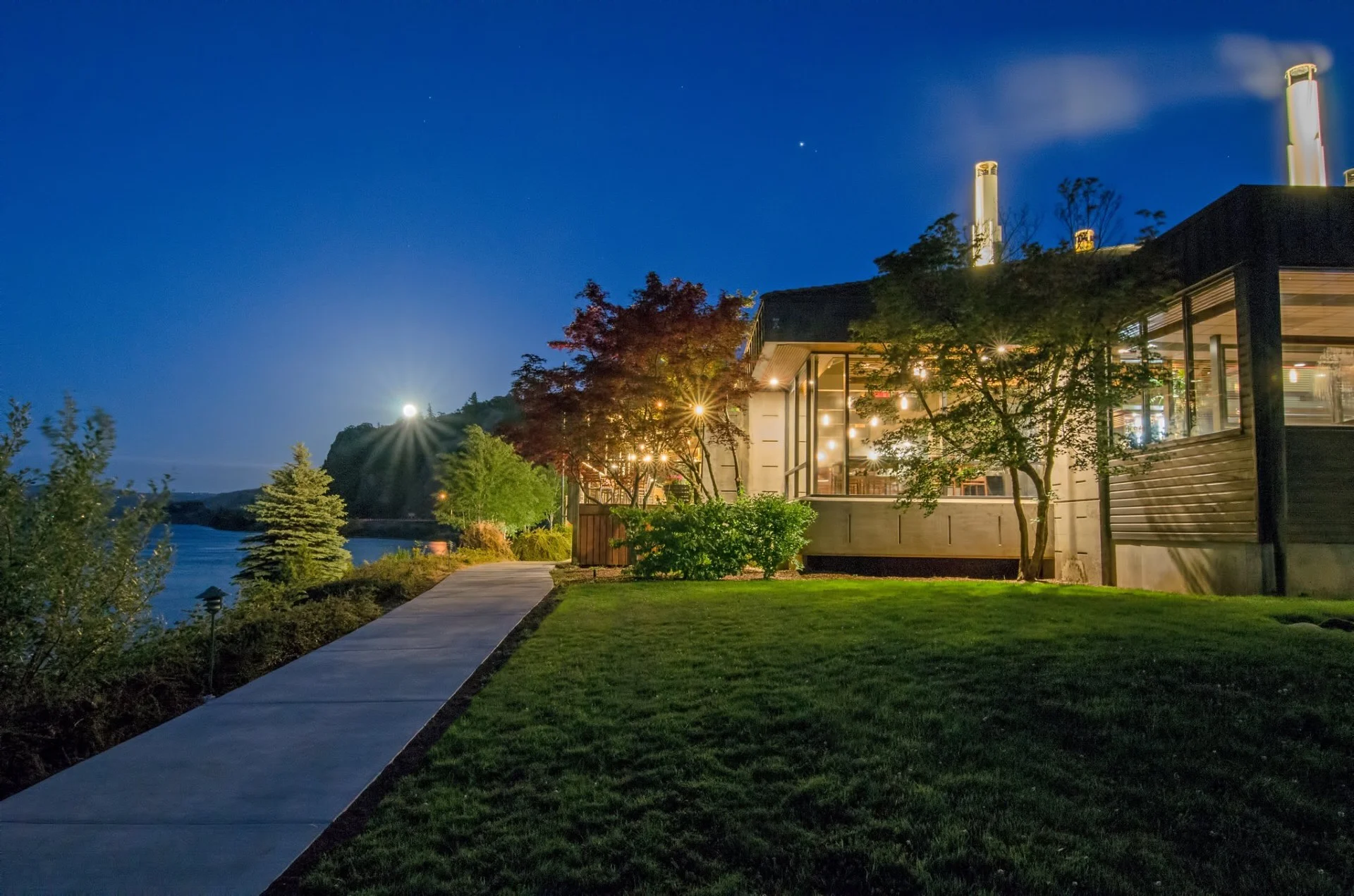 The inn lit warmly from within looks out over the river and a riverfront walking path at dusk.