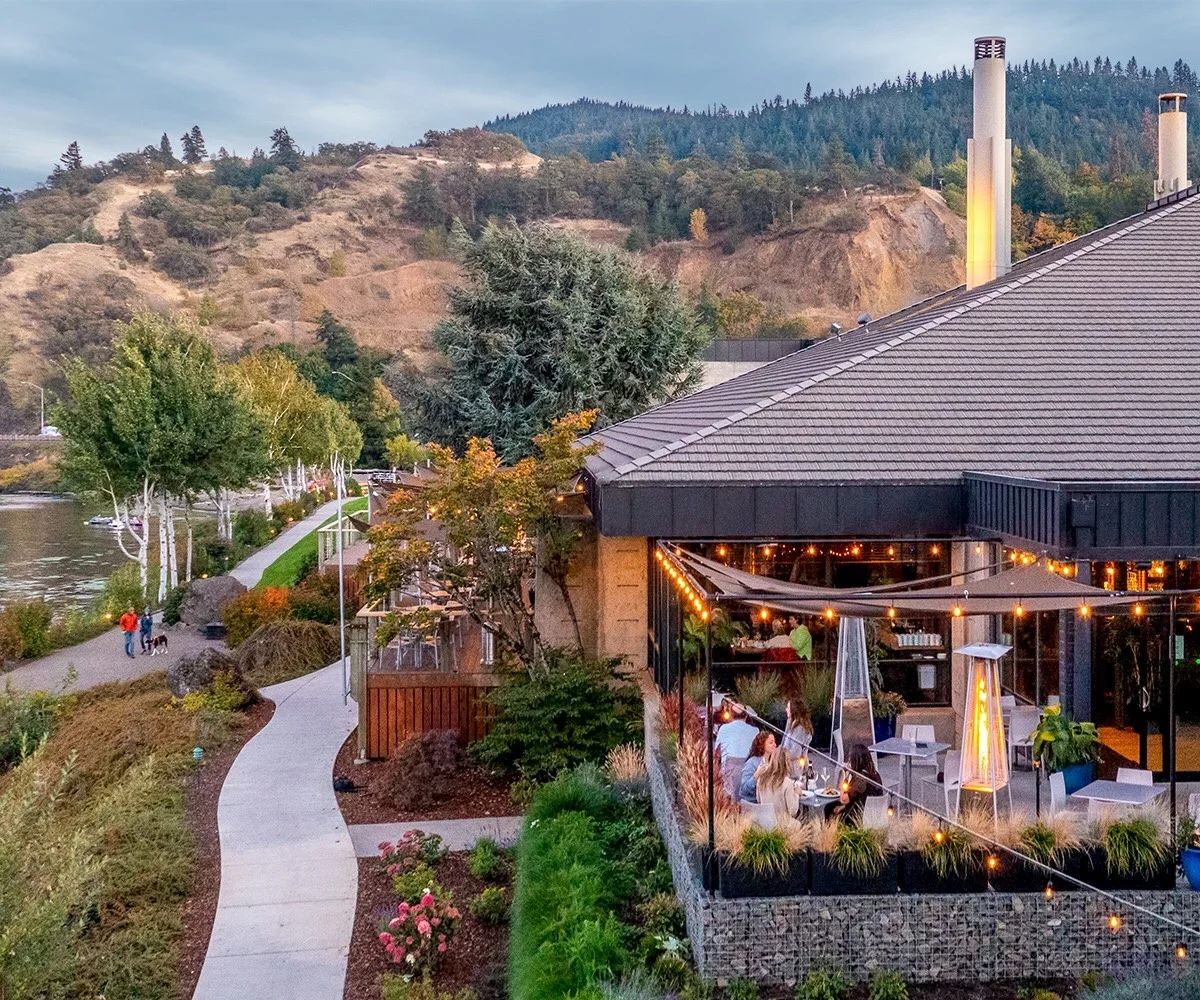 People dine at the Riverside Restaurant on an outdoor patio overlooking a riverfront walking path.