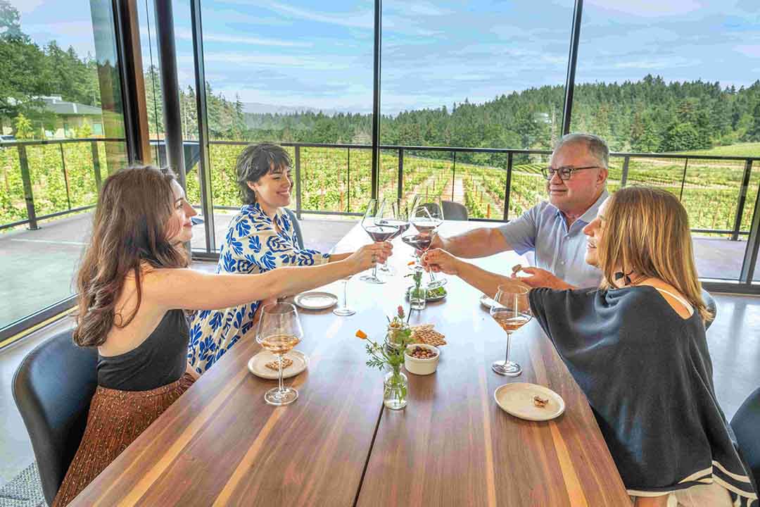 four people seated at a long wooden table raise wine glasses and cheers