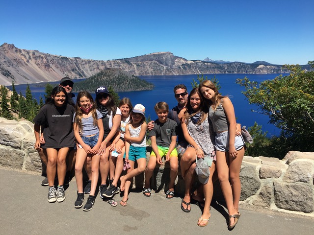 Tour group sits on a short stone wall in front of bright blue Crater Lake.