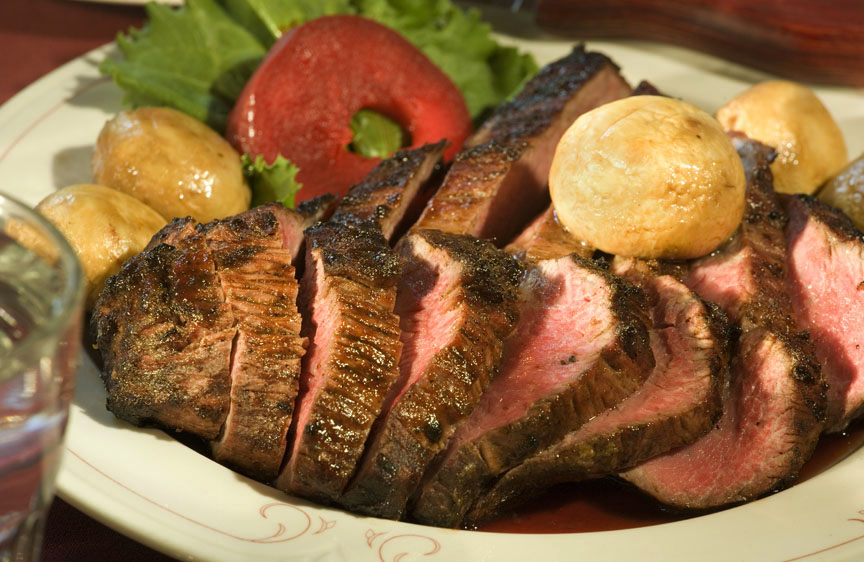 A white plate holds a sliced medium rare cooked steak with a couple golden potatoes and veggies to the side.