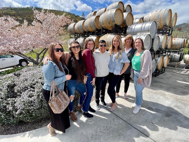 Group of seven women stand in front of wine barrels stacked vertically with cherry blossom tree beside them.