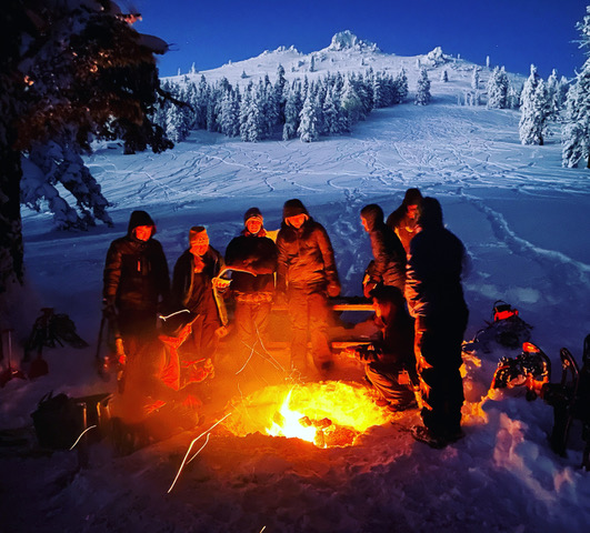 Tour group stands beside a roaring fire pit in the snow at dusk with a snowy hill lined with trees in the background.