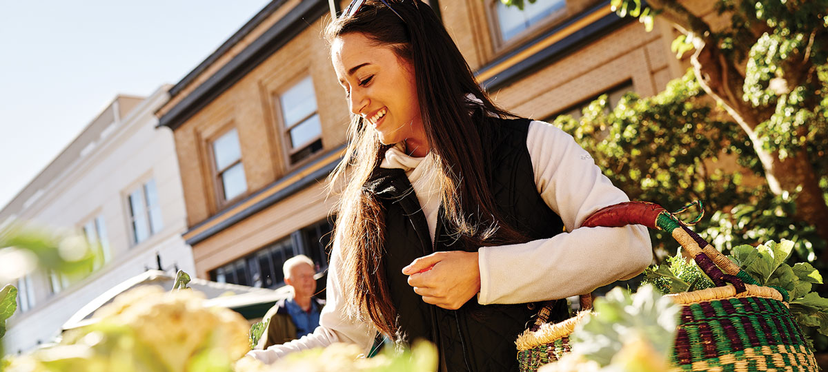 A smiling woman browses fresh produce at the Coos Bay Farmers Market, wicker basket in hand, with historic storefronts in the background.