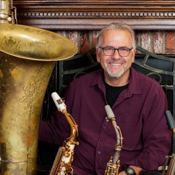 man wearing glasses, a maroon shirt sitting near a variety of saxaphones