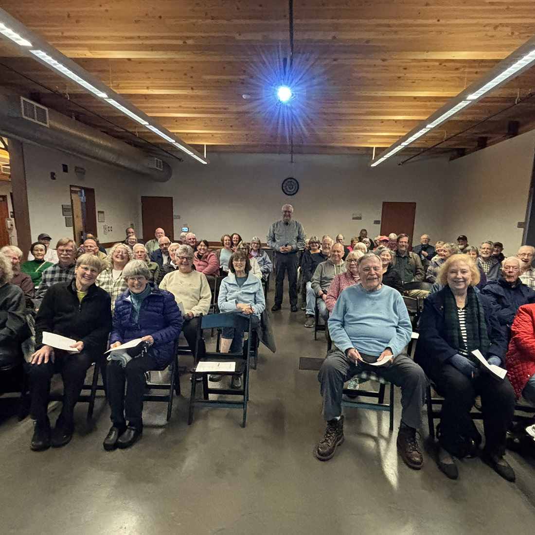 A large group of people seated in rows inside a meeting room, facing the camera, with a presenter standing at the back under a ceiling projector.