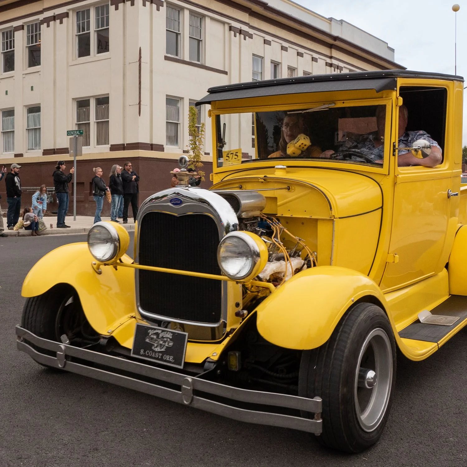 Bright yellow vintage hot rod drives along a downtown street during a classic car cruise, with spectators watching from the sidewalk.