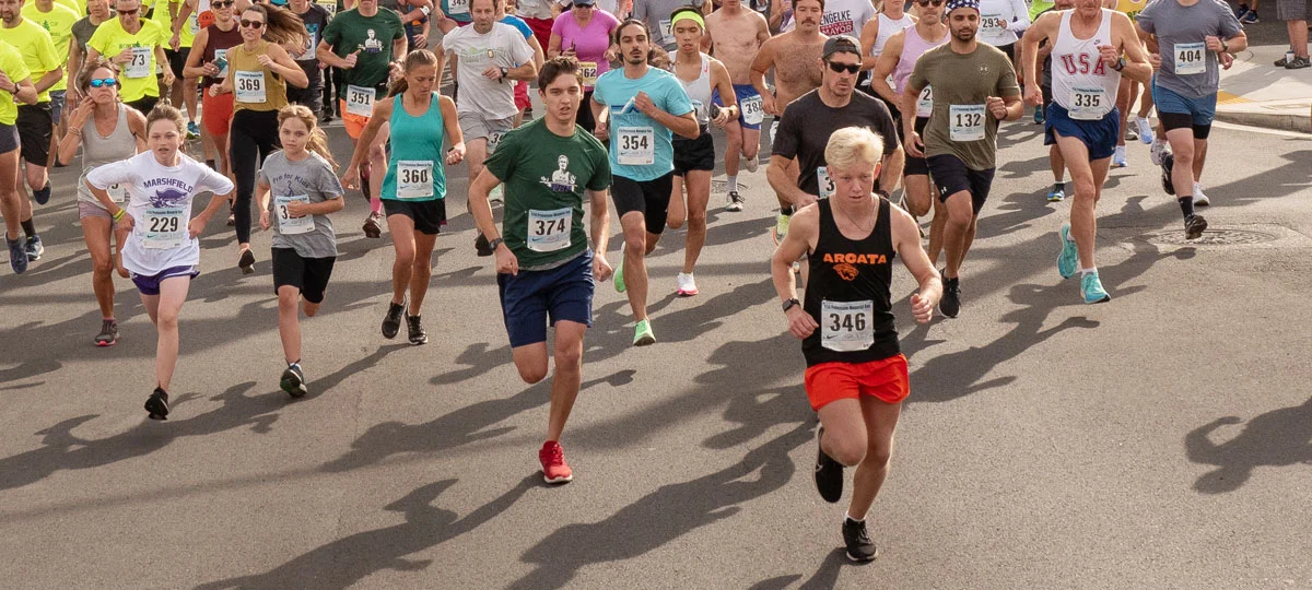 Participants of all ages run together during the Prefontaine Memorial Run on a city street in Coos Bay, with runners filling the course at the start of the race.