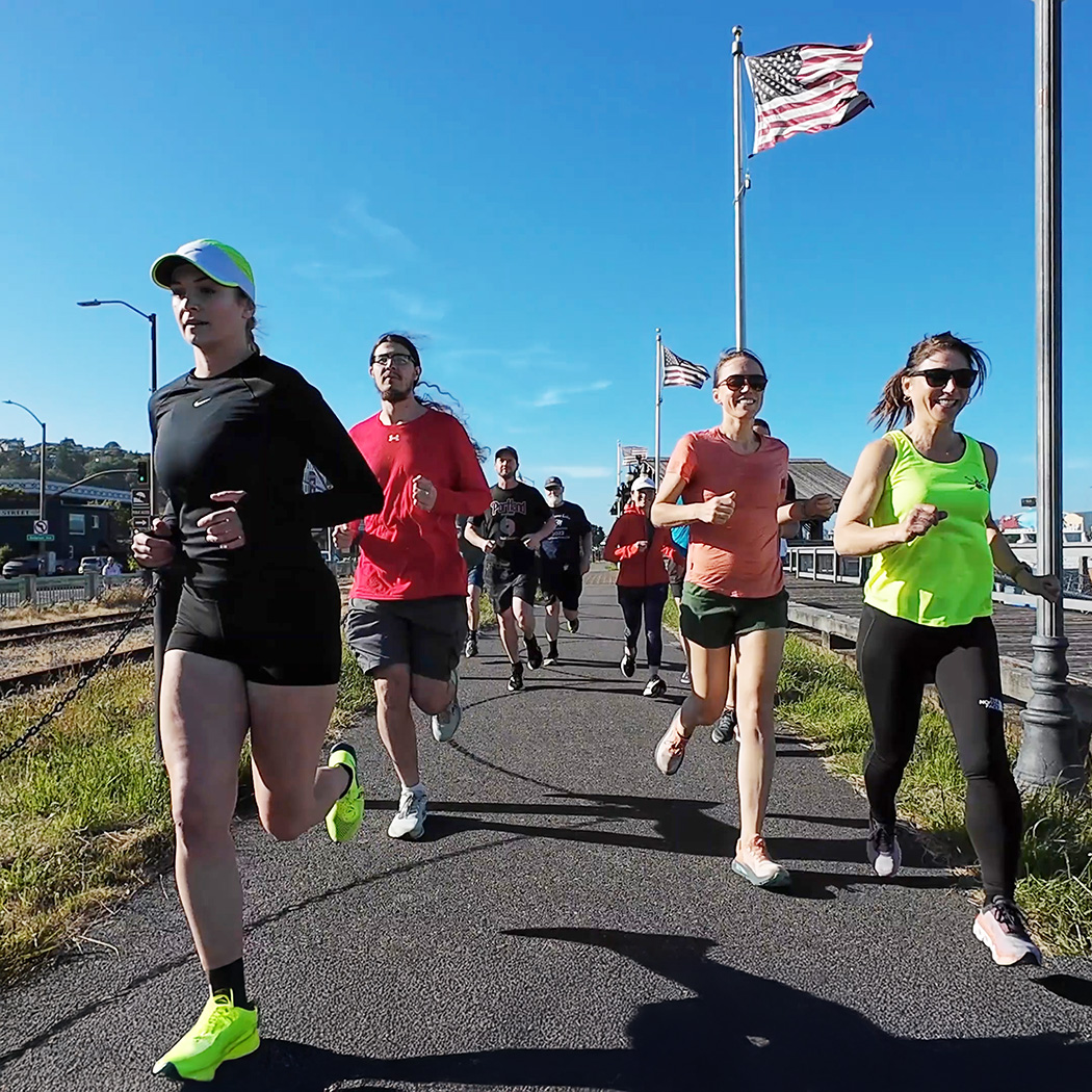 A group of runners jogs along a waterfront path beneath American flags on a sunny day. Two women in bright athletic tops lead the group while others follow on the paved trail beside the boardwalk.