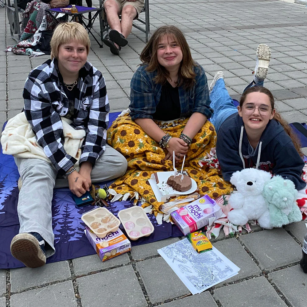 Three young people sit together on blankets with snacks, stuffed animals, and coloring pages while attending an outdoor movie night on a paved plaza.