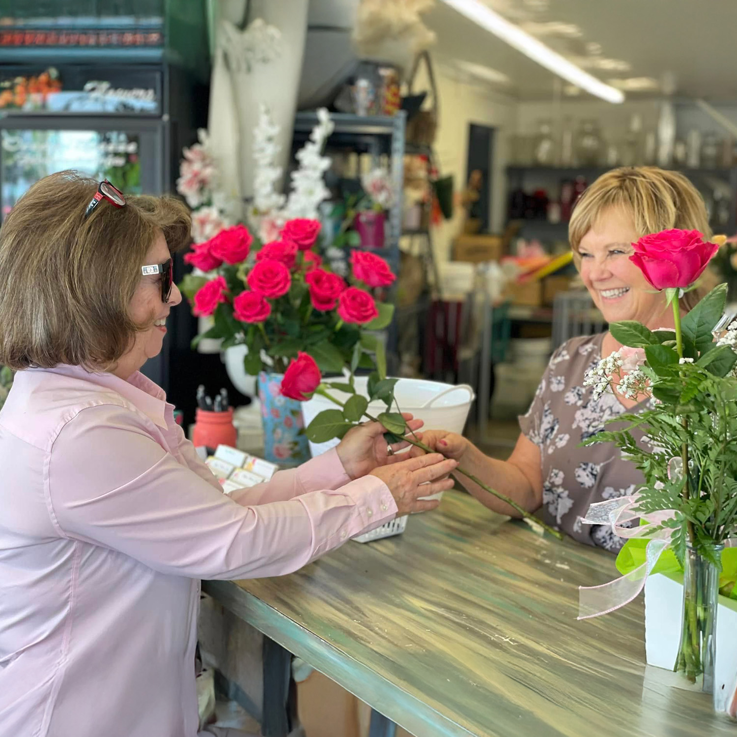 Customer and florist smiling as a single pink rose is handed across a flower shop counter, with bouquets and vases in the background.