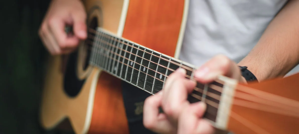 A close-up view of hands playing an acoustic guitar, with the strings and fretboard in sharp focus while the musician and background are softly blurred.
