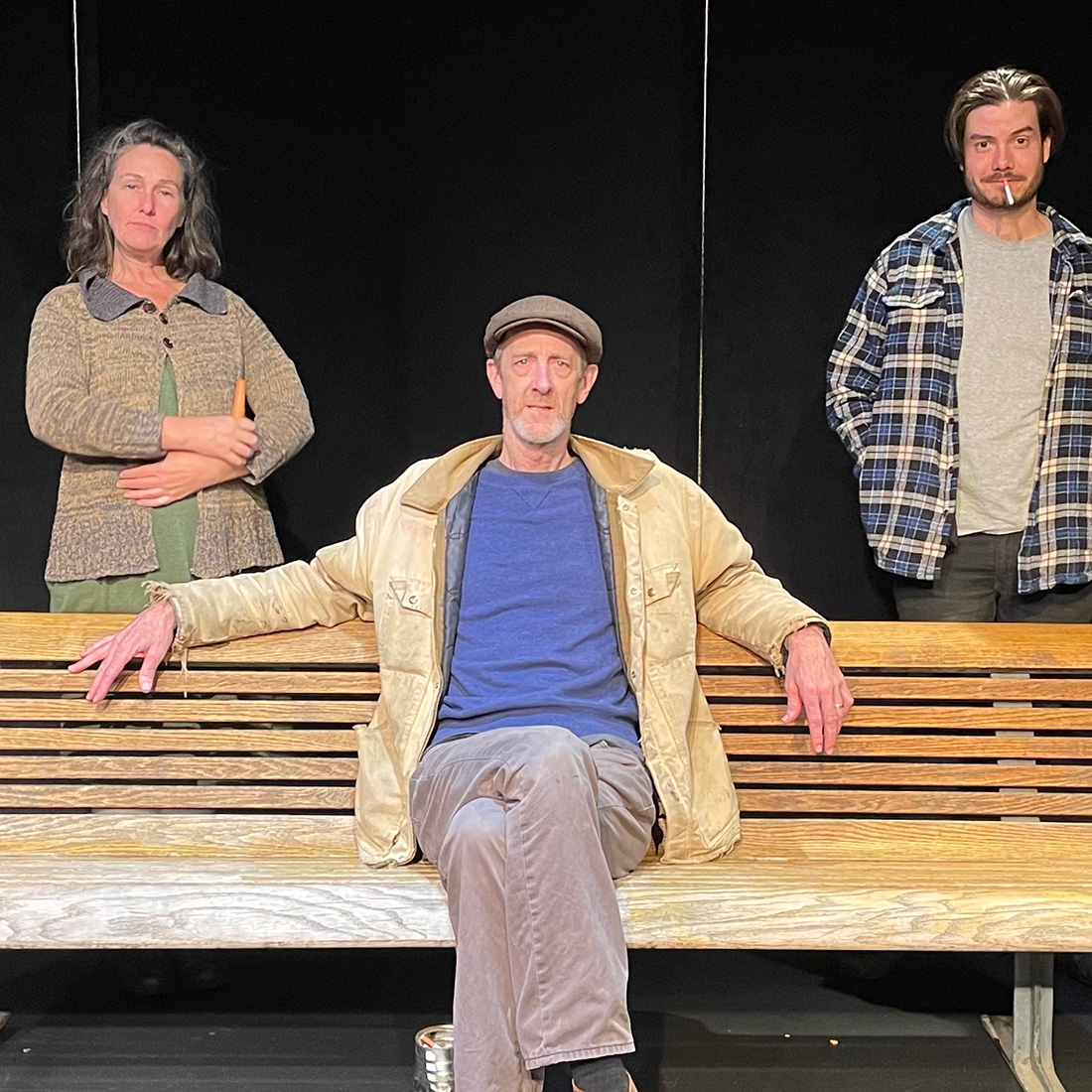 a wooden park bench with a man sitting on it.  A woman and and another man standing behind the bench.