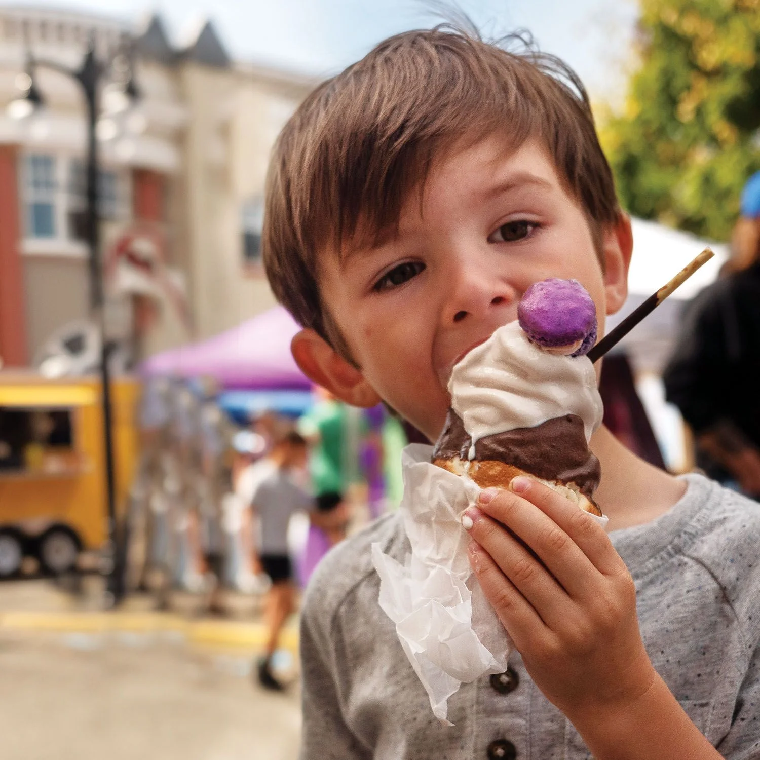 Child enjoying an ice cream cone topped with whipped cream and a purple macaron at an outdoor festival, with colorful vendor tents and a food truck blurred in the background.