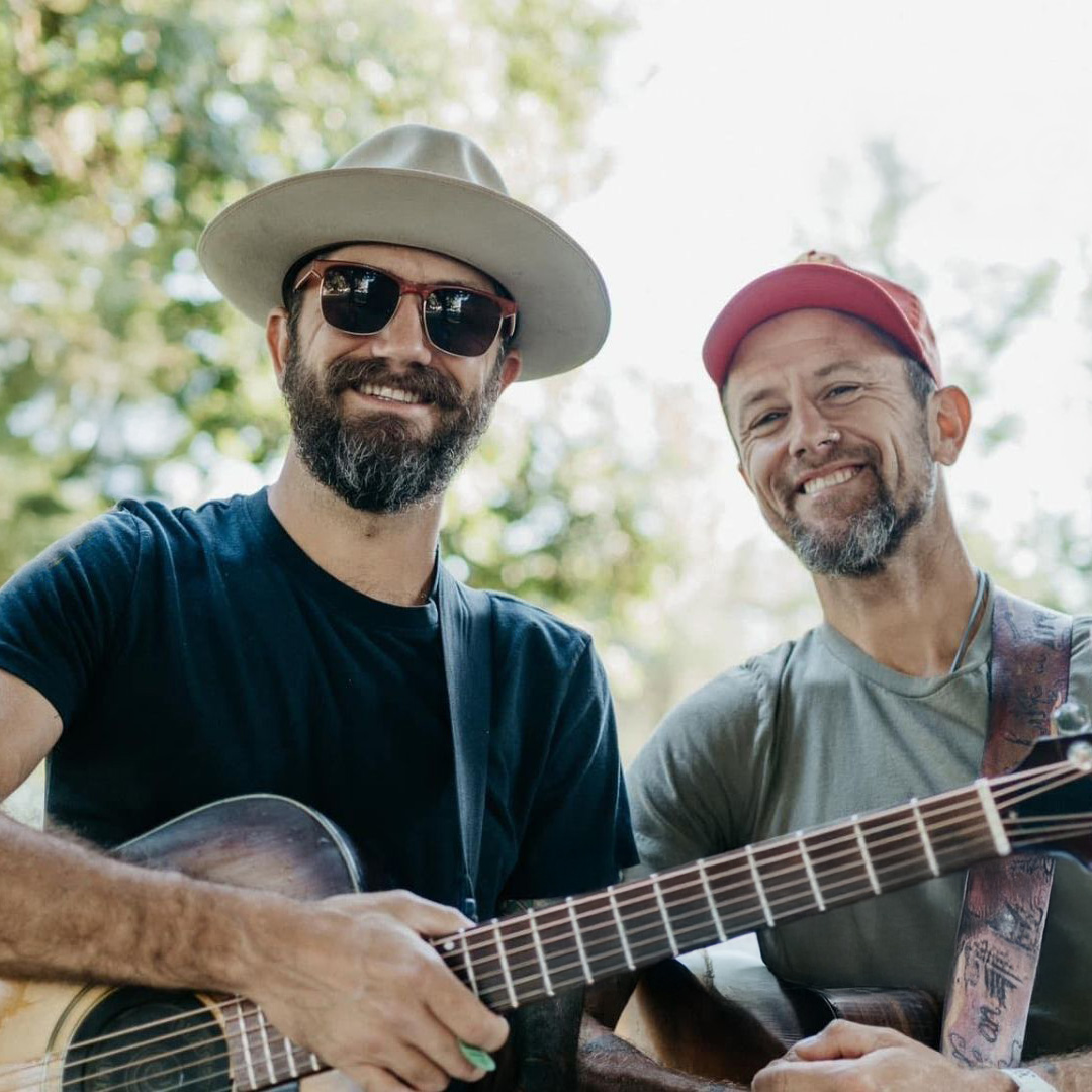 Two smiling men holding guitars outdoors, one wearing a wide-brim hat and sunglasses, the other in a red cap.