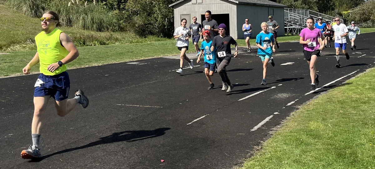 A runner leads the field during a community race on an outdoor track as participants of all ages follow behind, wearing numbered bibs on a sunny day.