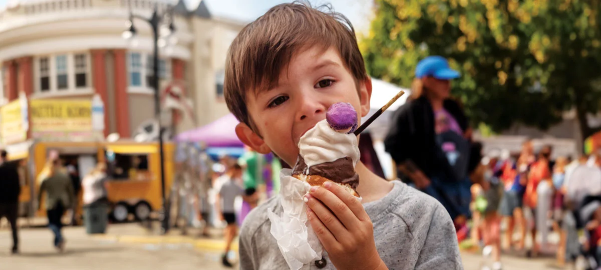 Child enjoying an ice cream cone topped with whipped cream and a purple macaron at an outdoor festival, with colorful vendor tents and a food truck blurred in the background.