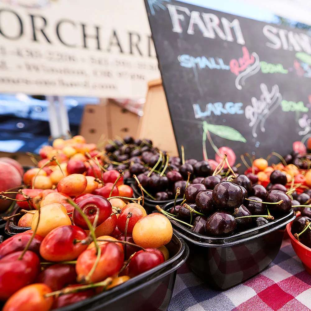 Baskets of red, yellow and dark cherries are displayed at a farmers market stand, with a colorful farm sign in the background.