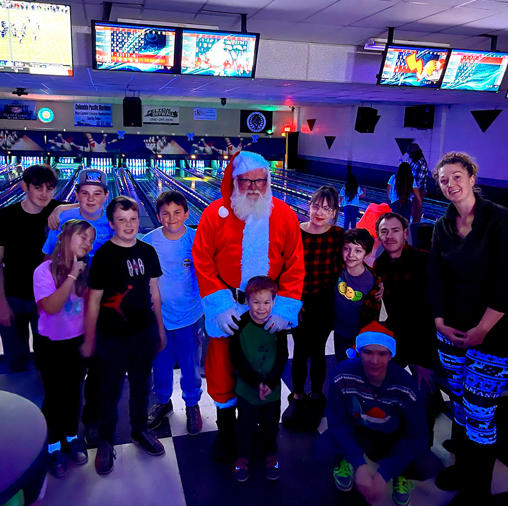 A man wearing a Santa Suit surrounded by young kids inside of a bowling alley