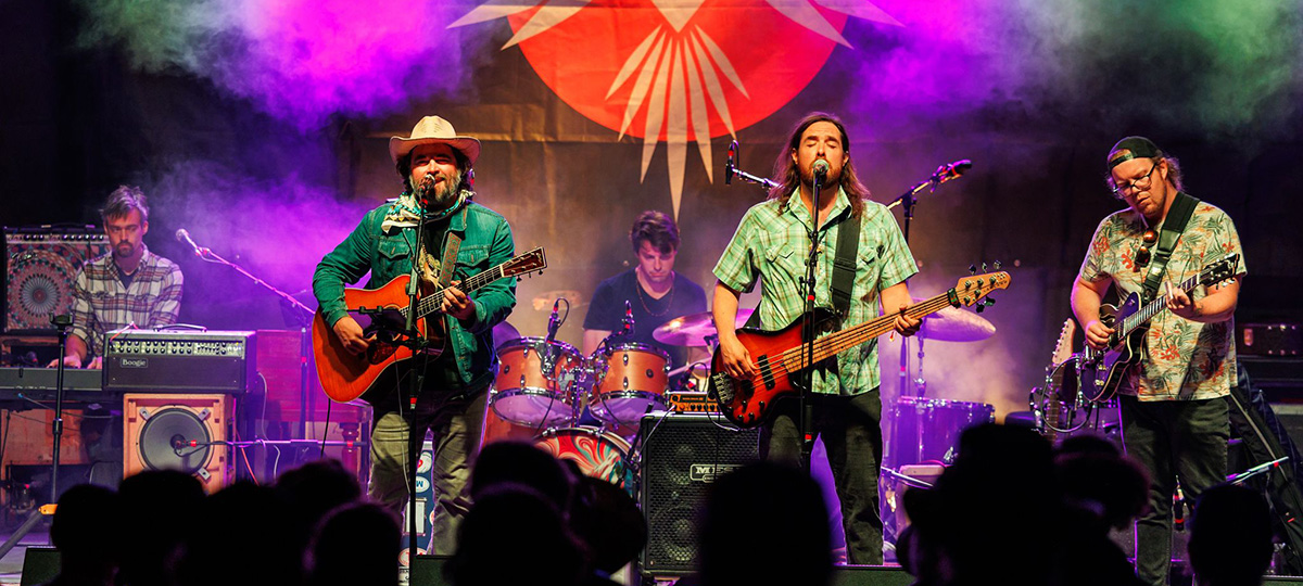 Band performing live onstage with colorful purple and pink lighting, featuring two musicians singing and playing guitar and bass in front of a drummer, with audience silhouettes visible in the foreground.