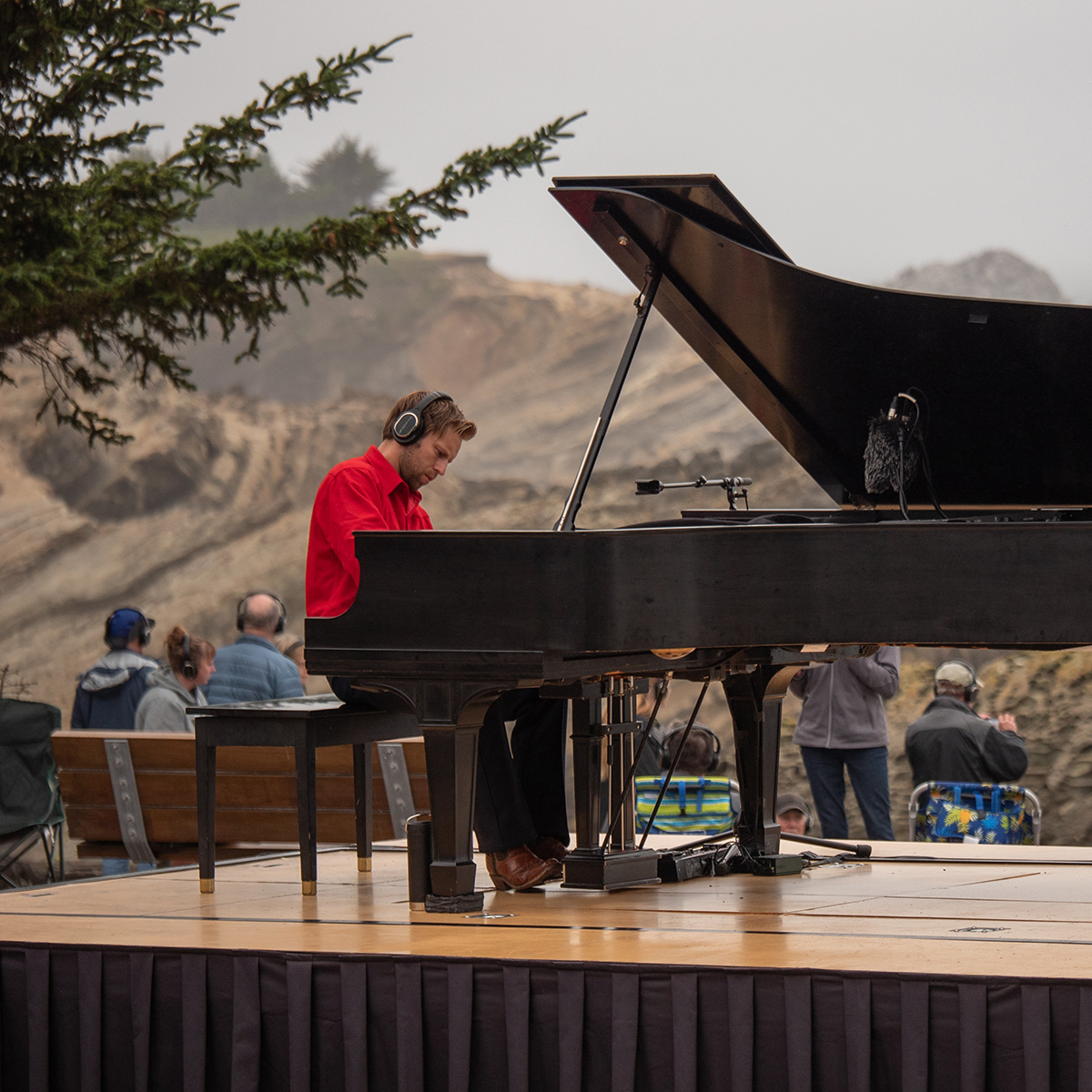Pianist in a red shirt wearing headphones plays a grand piano on an outdoor stage with rocky coastal cliffs in the background and seated audience nearby.
