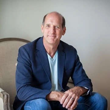 bald man in blue blazer and jeans sitting in a chair