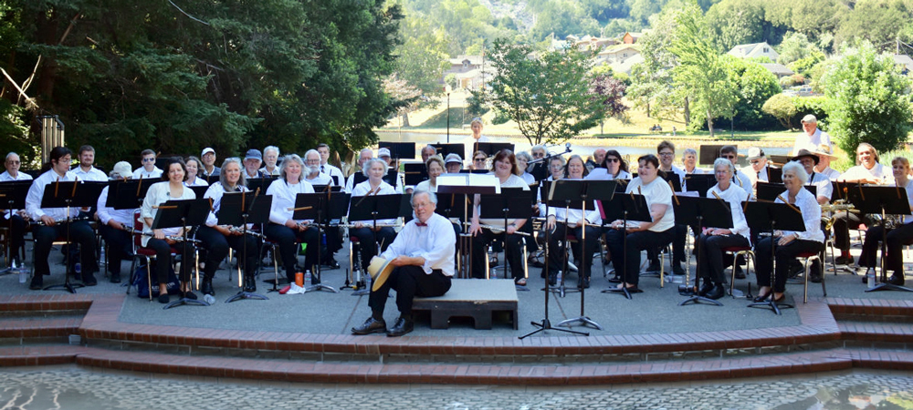 Community concert band members seated outdoors in a park amphitheater, performing with music stands and instruments before a wooded hillside and neighborhood backdrop.