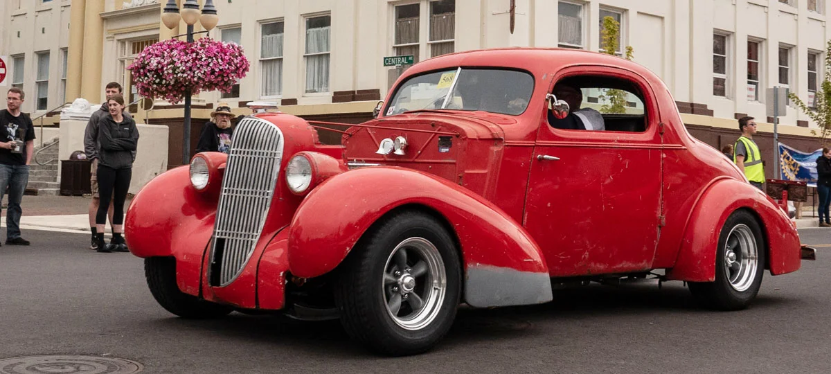 Red vintage coupe cruises through downtown Coos Bay during a classic car event, with spectators and hanging flower baskets lining the street.