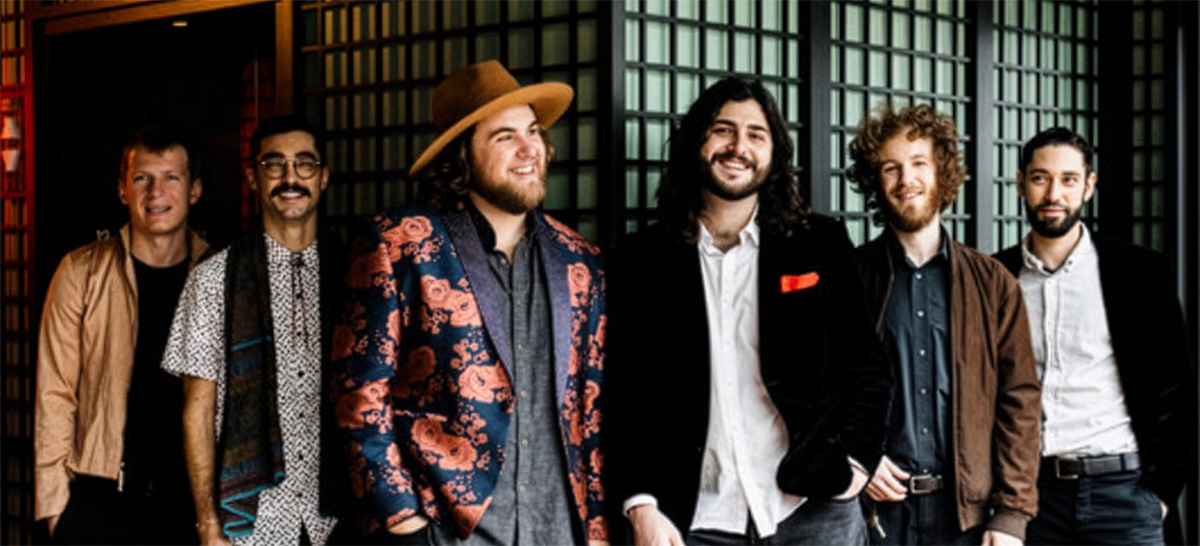 Six people standing together indoors on a black-and-gold checkered floor, wearing a mix of casual and dressy clothing, posing and smiling in front of decorative paneled walls.