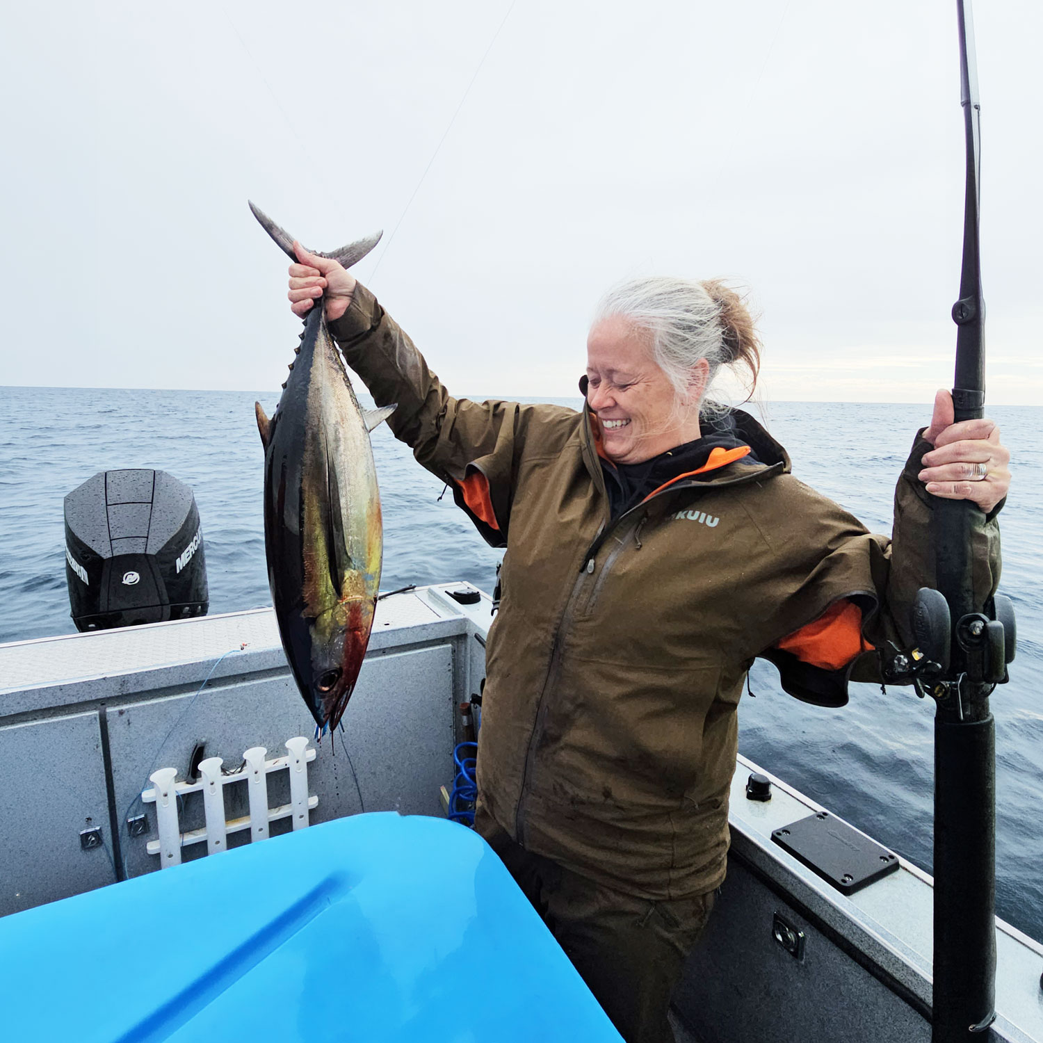 Woman on a fishing boat in open ocean waters, smiling while holding a freshly caught tuna in one hand and a fishing rod in the other. Overcast sky and calm sea in the background.