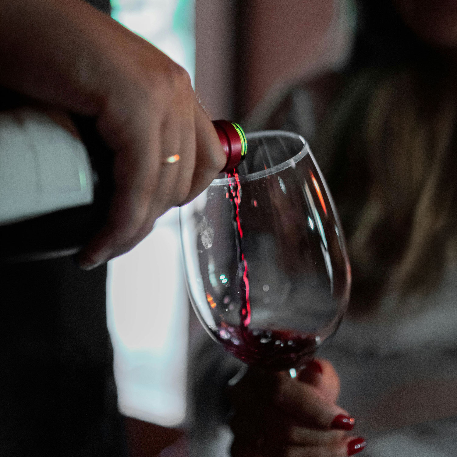 Close-up of red wine being poured from a bottle into a stemmed wine glass, with a person holding the glass. The background is softly blurred, emphasizing the pouring wine and creating a warm, intimate tasting atmosphere.