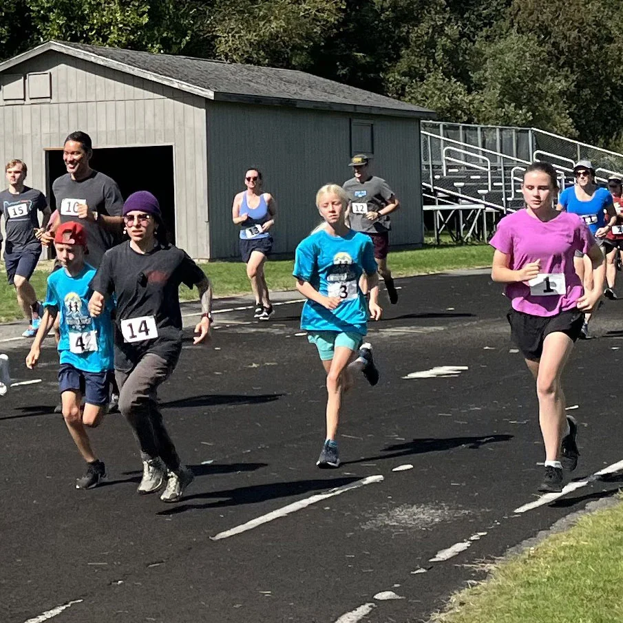 Runners of all ages compete on an outdoor track during a community race, with participants wearing numbered bibs as spectators and buildings appear in the background.