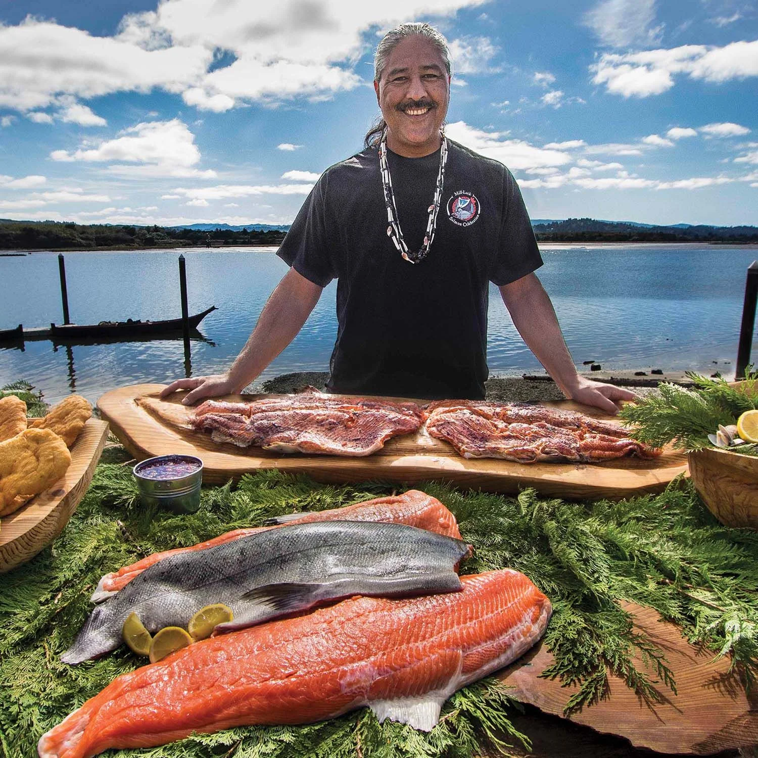 Person standing behind fresh salmon fillets and traditional foods beside a river, with canoes on the water and a blue sky in the background during a Native cultural celebration.
