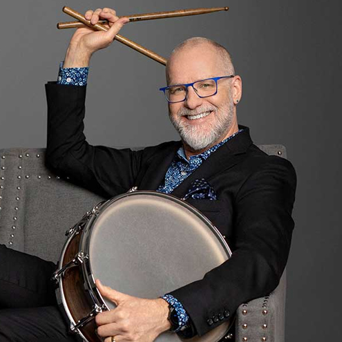 Person wearing glasses and a dark jacket smiles while seated in a chair, holding a drum and raising a pair of drumsticks against a gray background.