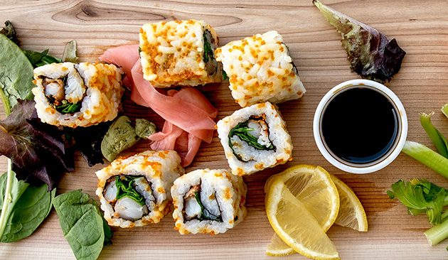 Sushi rolls with greens and tempura flakes served with soy sauce, lemon slices, wasabi, pickled ginger, and mixed greens on a wooden board.