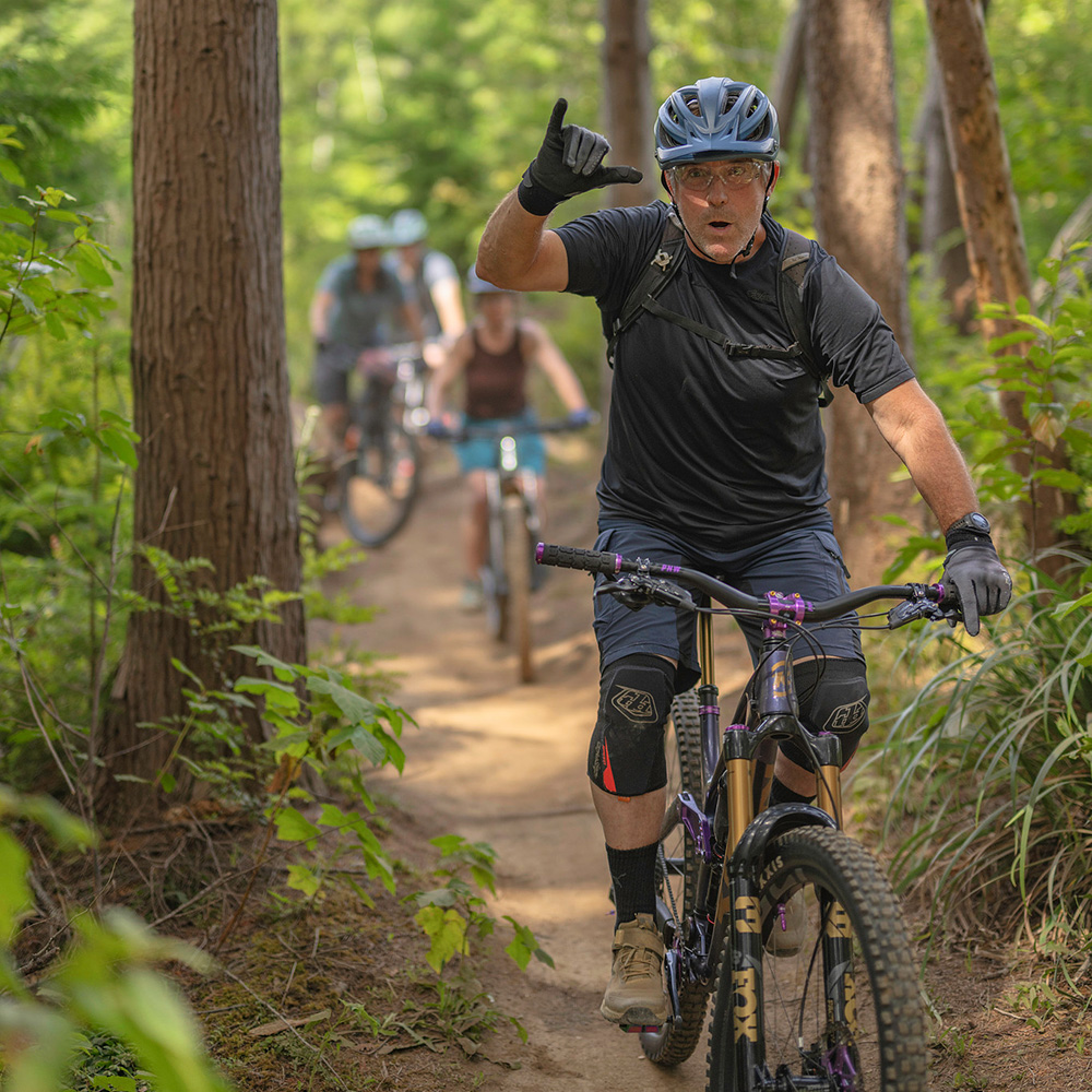 Man on a mountain bike riding on a trail in the woods followed by other riders in the distance