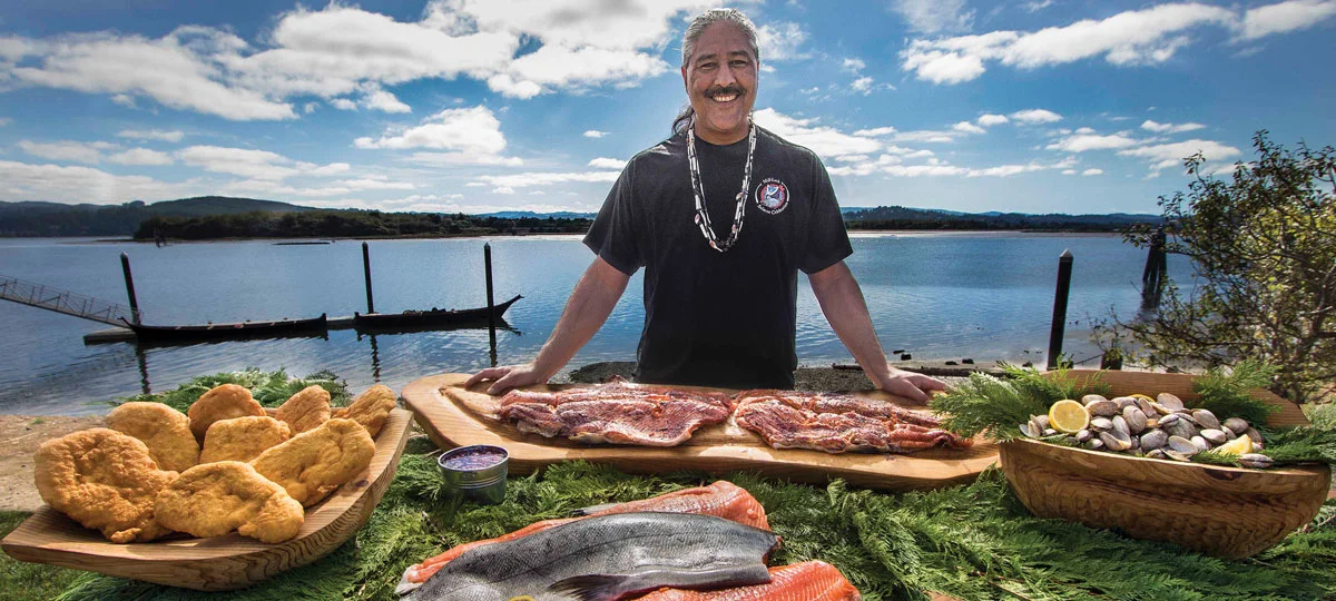 Person standing behind fresh salmon fillets and traditional foods beside a river, with canoes on the water and a blue sky in the background during a Native cultural celebration.