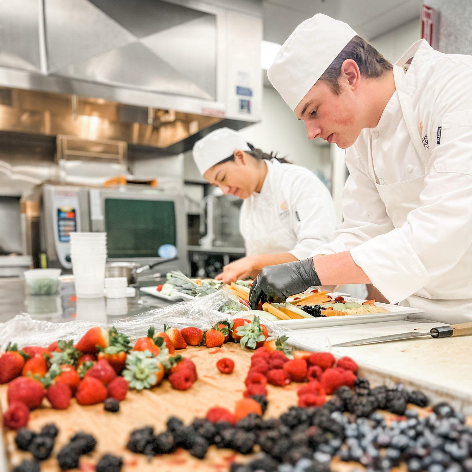 Two chefs in a commercial kitchen carefully preparing fruit trays with fresh berries on the counter.
