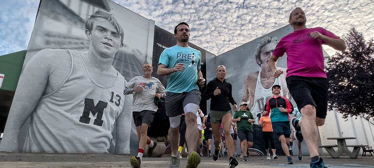 Runners jog past large black-and-white murals of track athletes on an urban street. Participants of varying ages wear colorful athletic clothing beneath a sky filled with scattered clouds.