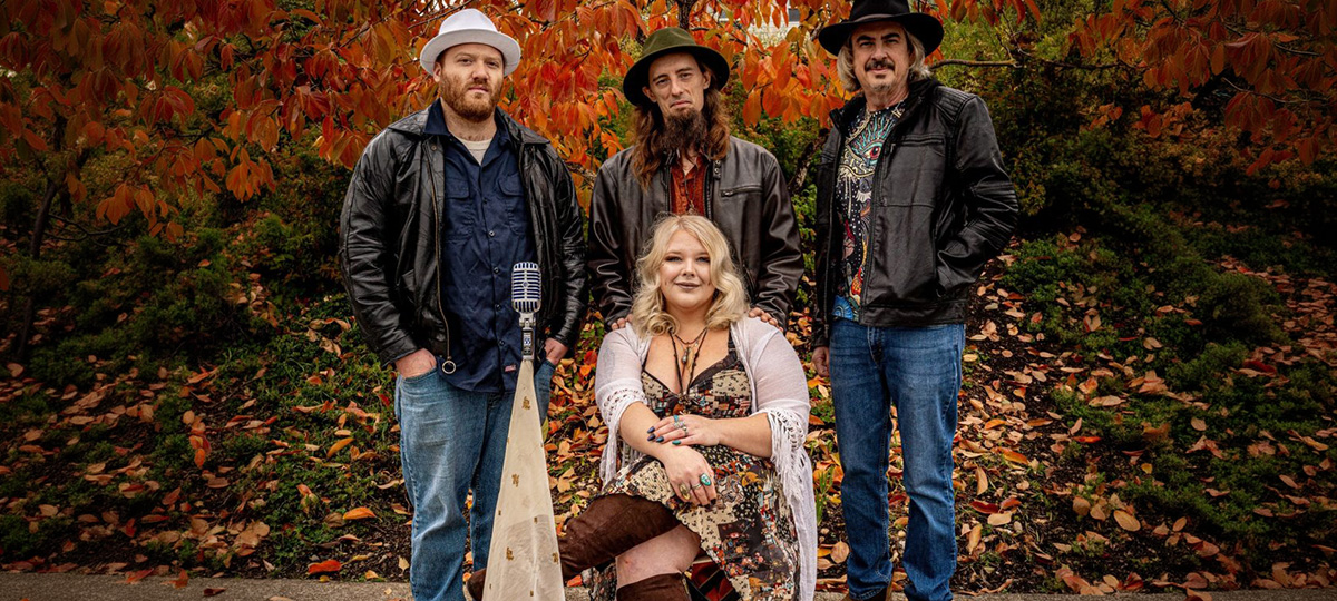 Four musicians posing outdoors in front of autumn foliage, with one seated and three standing beside musical equipment including a microphone.