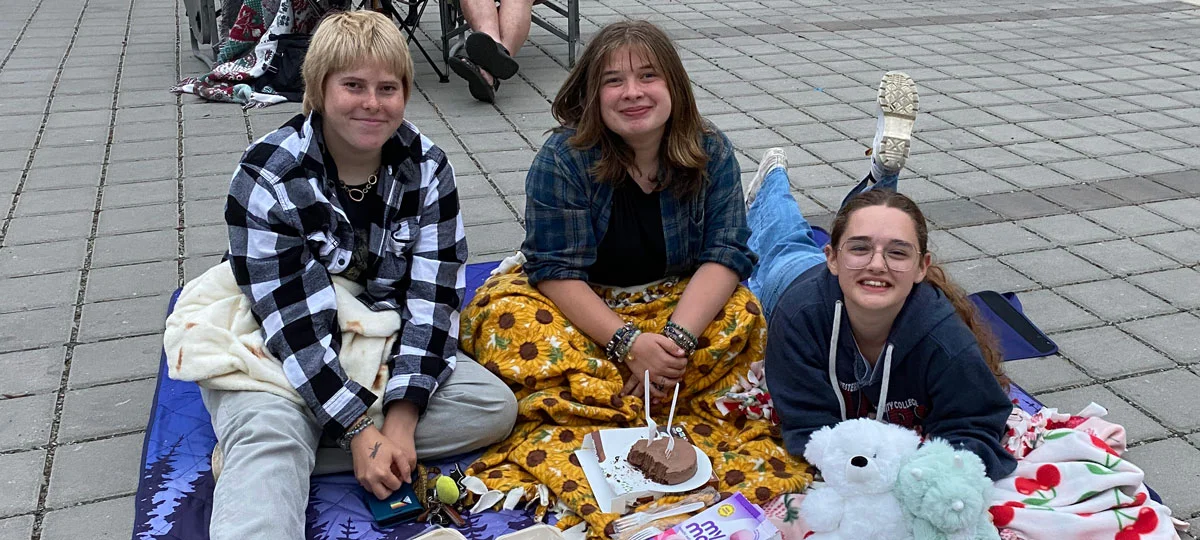 Three young people sit together on blankets with snacks, stuffed animals, and coloring pages while attending an outdoor movie night on a paved plaza.