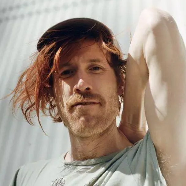 Person with windswept reddish hair and a light-colored shirt looks toward the camera with one arm raised behind their head against a softly lit background.