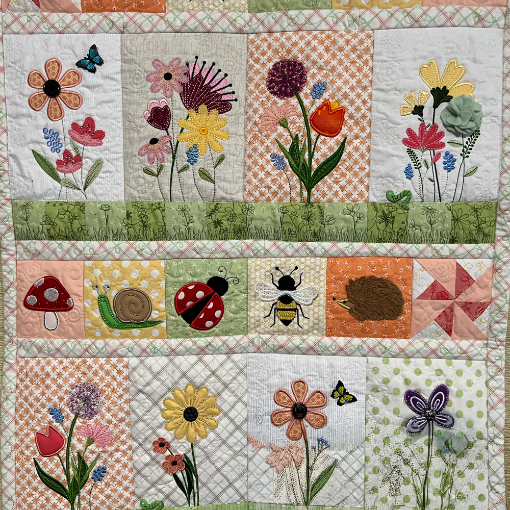 Colorful handmade quilt featuring embroidered flowers, butterflies, mushrooms, a snail, ladybug, bee, and hedgehog. Pastel fabrics, decorative stitching, and patchwork blocks create a whimsical garden scene.
