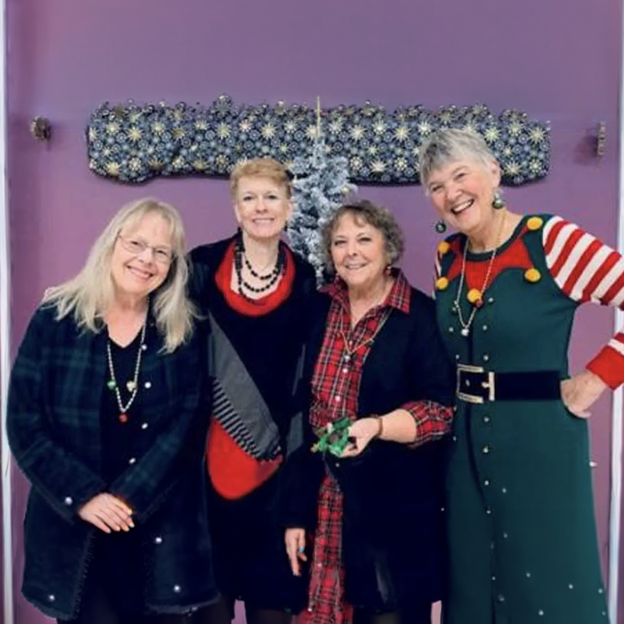 Four women stand smiling together indoors in festive holiday attire, posing in front of a purple wall with decorative artwork and a small silver tree.