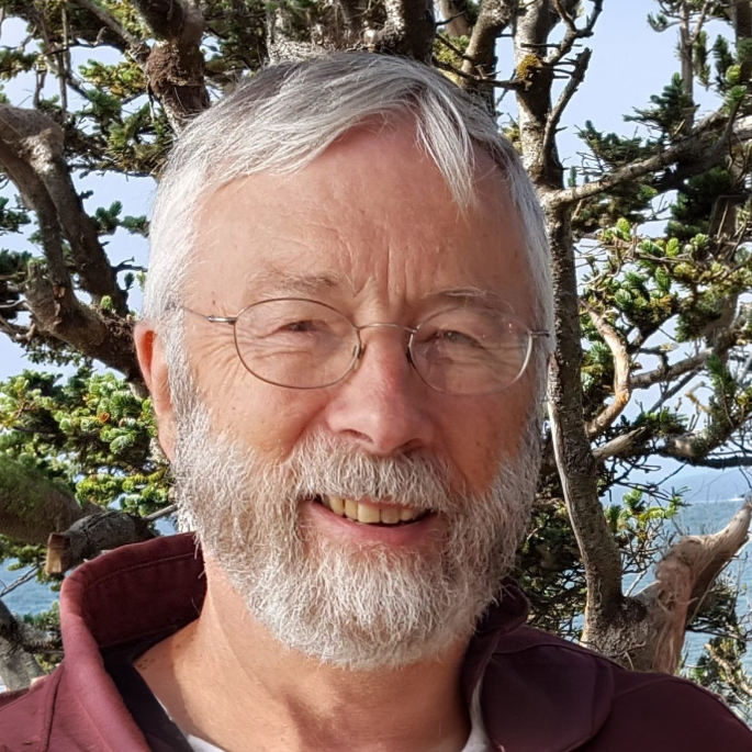 Portrait of a smiling man with a gray beard and glasses standing outdoors. Trees and coastal vegetation frame the background, with a glimpse of water visible beyond the branches.
