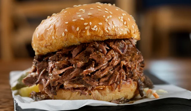 Pulled pork sandwich from Yoda's Hut piled high with slow-cooked barbecue pork on a sesame seed bun, served on deli paper with pickles.