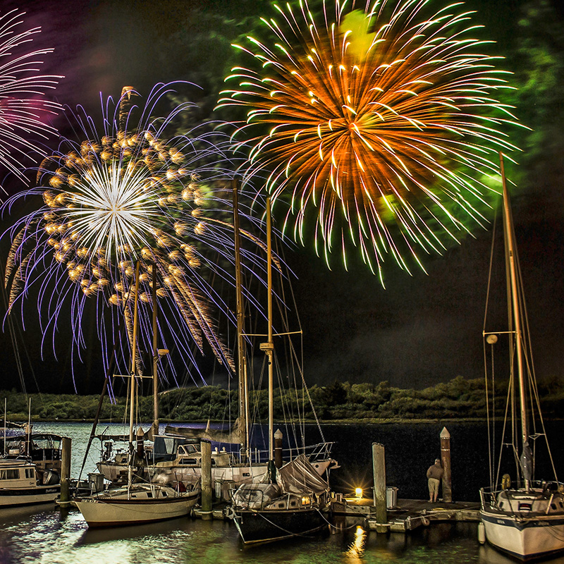 the night sky lit up by fireworks over a marina filled with sailboats with sand dunes in the distance