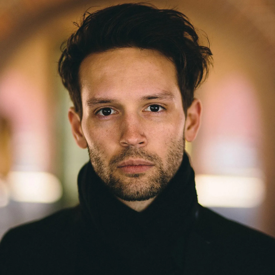 A close-up portrait of a person with dark, styled hair and light facial stubble, wearing a black high-collared coat, looking directly at the camera with a neutral expression against a blurred background.