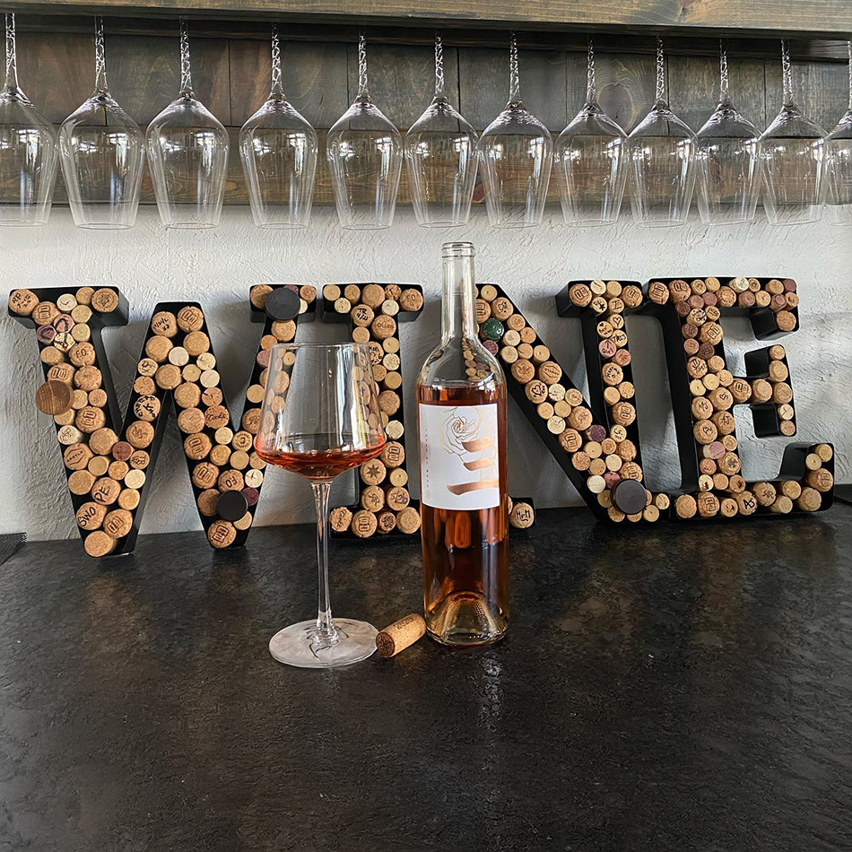 A bottle and glass of rosé wine in front of cork-decorated letters spelling “WINE,” with glasses hanging above.
