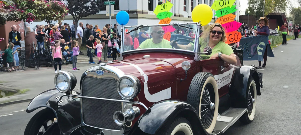 Vintage red convertible decorated with colorful balloons and "Oh, the Places You'll Go!" signs drives in a community parade as two smiling riders wave and spectators line the street.